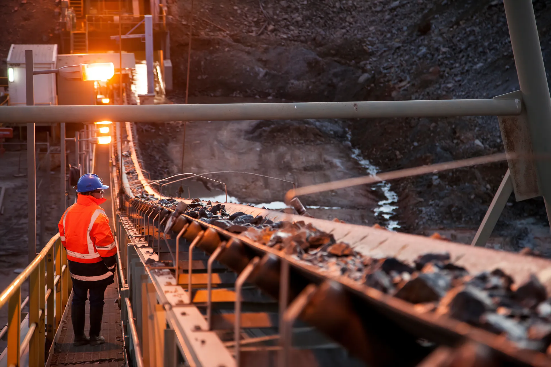 Mining conveyor carrying ore at dusk with plant lights in the background